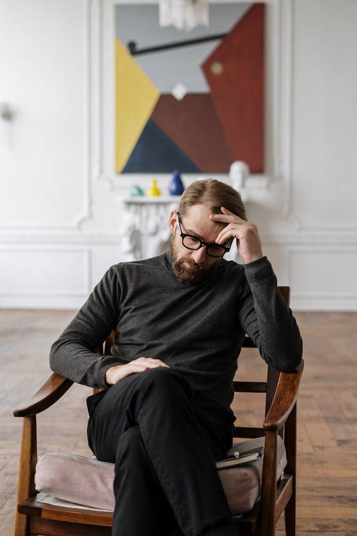 A man sits pensively in a chair, surrounded by abstract art in a stylish room.